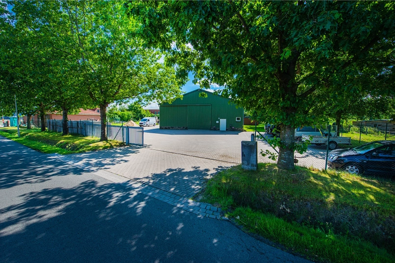 Zu sehen ist die gepflasterte Straßenzufahrt zum Gebäude bzw. der dunkelgrünen Halle des Bauhofes in Ahlerstedt. Umringt wird das Gelände vom einen Zaun sowie hohen Bäumen, die große  Schatten werfen.