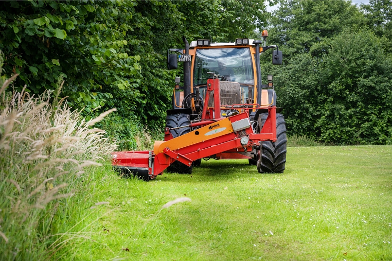 Zu sehen ist auf einer Rasenfläche ein Traktor mit Frontmährwerk beim Einsatz, welcher von einem Mitarbeiter des Bauhofes in Ahlerstedt bedient wird. Umrandet wird die Rasenfläche von Bäumen.