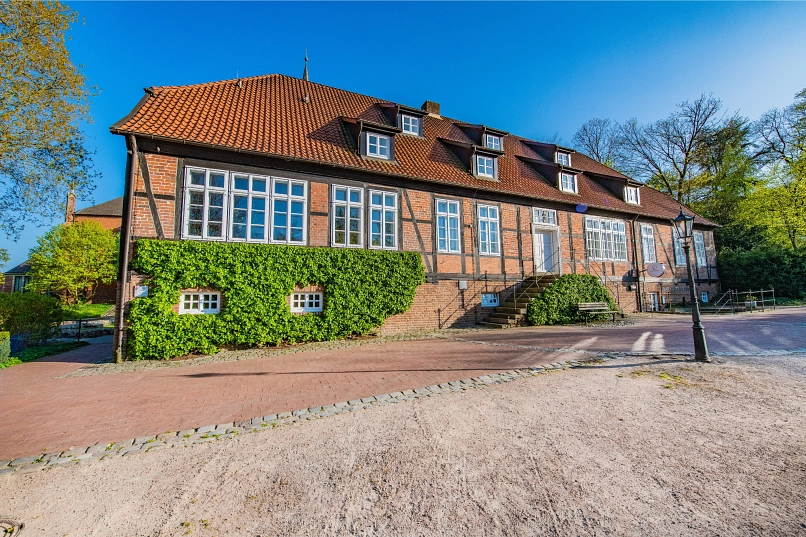 Der Amtshof Harsefeld mit bewachsener Wand am gepflasterten vor blauem Himmel und grünen Bäumen. Im Hintergrund ist die Kirche teilweise zu sehen.