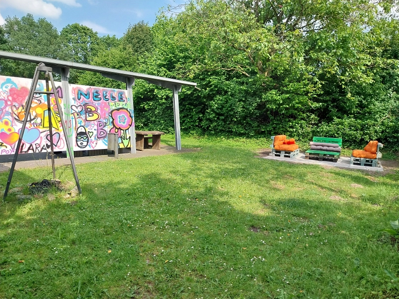 Eine Grünfläche mit Schwenkgrill und Paletten Sofa Ecke, ebenso ist eine bunte Graffitiwand zusehen.