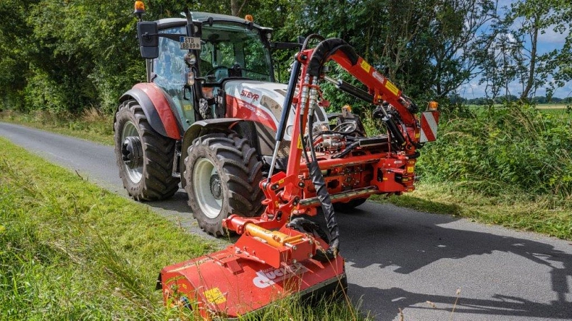 Zu sehen ist ein roter STEYR Traktor mit einem Frontmähwerk, welcher auf einer schmalen Straße den Seitenstreifen mäht. Gefahren wird der Traktor von einem Mitarbeiter des Bauhofes in Bargstedt.