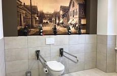Barrierefreie WC-Anlage Museum Harsefeld Sanitärraum mit barrierefreier Toilette inklusive Haltegriffe und Notfall-Zugschnur. Im Hintergrund an der Wand ein historisches Bild von der Markstraße Harsefeld