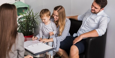 Zu sehen ist eine Familie auf dem Sofa, die auf ein Blatt Papier schauen. der Familie gegenüber sitzt eine Frau, die sie berät.