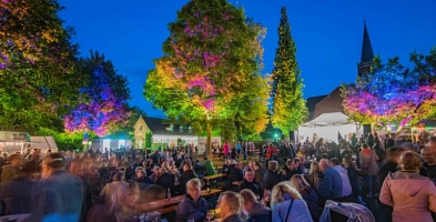 Der Klosterpark mit vielen Menschen zum Bierzauber. Die Menschen sitzen auf Bierzeltgarnituren, im Hintergund ist eine Bühne mit Livemusik zu sehen und die Bäume werden bunt angestrahlt.