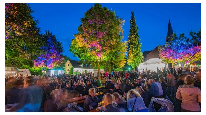 Der Klosterpark mit vielen Menschen zum Bierzauber. Die Menschen sitzen auf Bierzeltgarnituren, im Hintergund ist eine Bühne mit Livemusik zu sehen und die Bäume werden bunt angestrahlt.