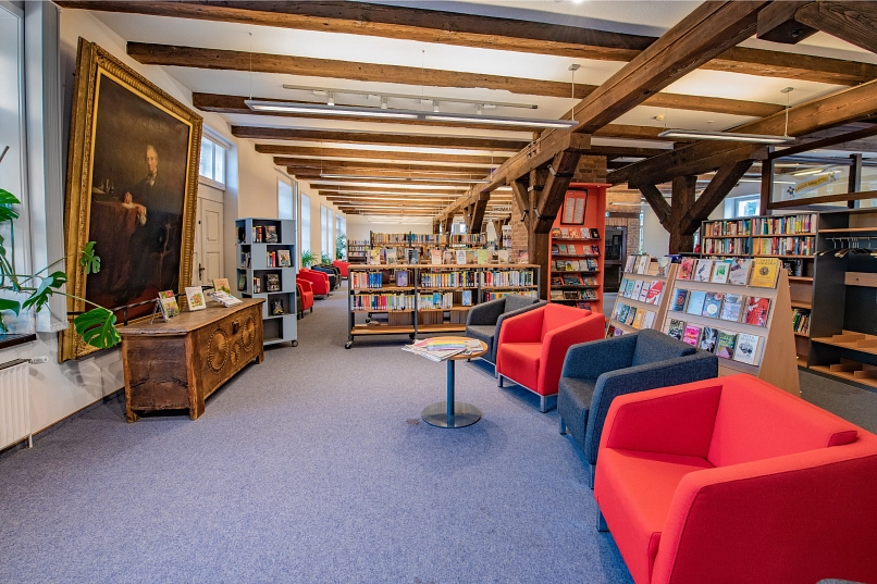 Vier Sessel vor einer Truhe hinter der ein großes Gemälde von Friedrich Huth an der Wand hängt. Hinter und neben den Sesseln befinden sich Bücherregale.