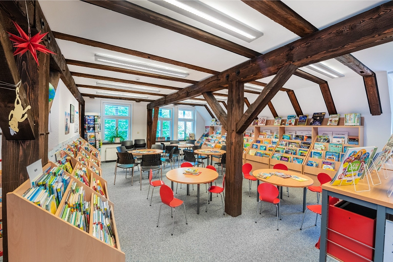 Sachbücher für Kinder in Regalen im Obergeschoss der Bücherei. Im Vordergrund hängen an einem Holzbalken Basteleien. In der Mitte des Raums sind 5 Tische mit je 5 Stühlen aufgestellt.
