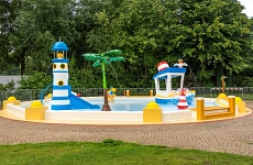 Zu sehen ist das bunte Kinderplanschbecken mit einem Leuchtturm, einer Palme und einem Boot, die herausstechen und aus denen Wasser spritzt. Weitere kleinere Wassertiere befinden sich im Becken.
