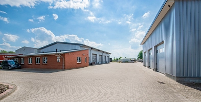 Zu sehen ist eine lange gepflasterte Hofeinfahrt. An den beiden Seiten sieht man jeweils Gewerbehallen mit drei großen Rolltoren. Vor der linken Halle ist zudem ein Büroanbau mit parkenden Auto zu sehen.
