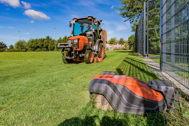 Zu sehen ist auf die Rasenfläche eines Sportplatzes samt Zaunumrisse im rechten Bereich. Im Vordergrund befindet sich ein Mähroboter im Einsatz. Dahinter wird von einem Mitarbeiter des Bauhofes ein Kommunaltraktor gefahren.