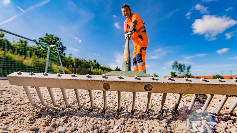 Zu sehen ist im Vordergrund eine Holzharke, welche durch eine Sandfläche mit Rasen gezogen wird. Unscharf dahinter erkennt man den Mitarbeiter des Bauhofes in Harsefeld, welcher die Harke hält. Ebenfalls sichtbar ist im linken Hintergrund ein Fussballtor.