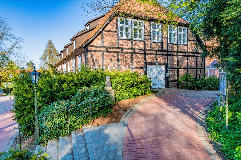 Eingang der Friedrich-Huth-Bücherei mit Schriftzug "Friedrich-Huth-Bücherei" neben der weißen Eingangstür des Amtshofs. Davor steht an einer Treppe die Büste von Friedrich Huth.