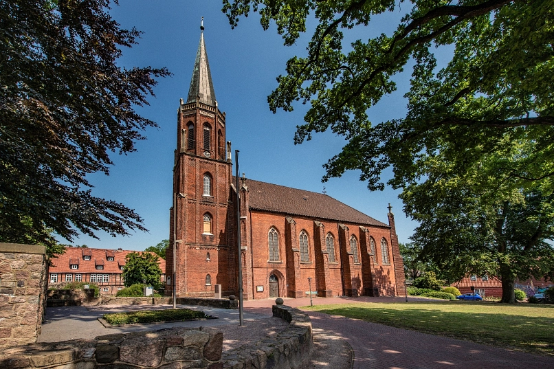 Ansicht einer Backsteinkirche mit linksseitigem Kirchturm, im Hintergrund historische Gebäude
