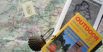 Landkarte mit darauf abgelegtem Wanderbuch, Pilgerflyer, einem Pilgerstempel und einer Jakobsmuschel