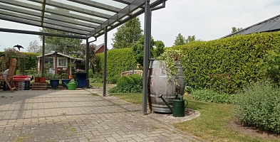 Im Vordergrung sieht man ein hölzernes Regenfass, das an einem Carportdach angeschlossen ist. Im Hintergrund ist ein Gartenhaus, Blumenbeete und auf der rechten Seite eine Kirschlorbeerhecke zu sehen.