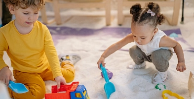 Auf dem Bild zu sehen sind zwei kleine Kinder, die ca. 1,5 Jahre alt sind und mit Baufahrzeugen im Sand spielen.