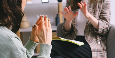 Zwei Frauen sitzen sich gegenüber und unterhalten sich. Eine hält ihre Hände zusammen, während die Frau im Hintergrund einen Stift in der Hand hält und ein Buch zum Schreiben auf dem Schoß hat.