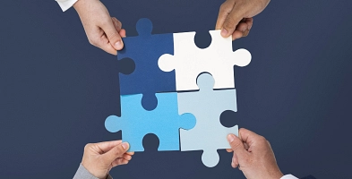 4 Hände, die jeweils ein Puzzleteil halten und in der Mitte zusammensetzen.
