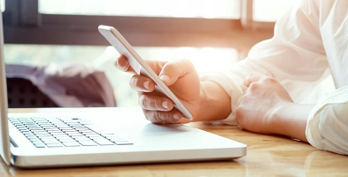 Eine Person benutzt ein Smartphone während ein Laptop vor ihr auf einem Tisch steht.
