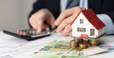 Ein Miniaturhaus steht auf mehreren Euro-Münzen und Scheinen. Darunter ist ein Graph zu sehen und eine Person im Hintergrund, die einen Anzug trägt und am Taschenrechner Berechnungen durchführt.
