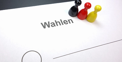 Drei Spielfiguren in den Farben Schwarz, Rot und Gelb auf einem Wahlschein mit der Aufschrift Wahlen.