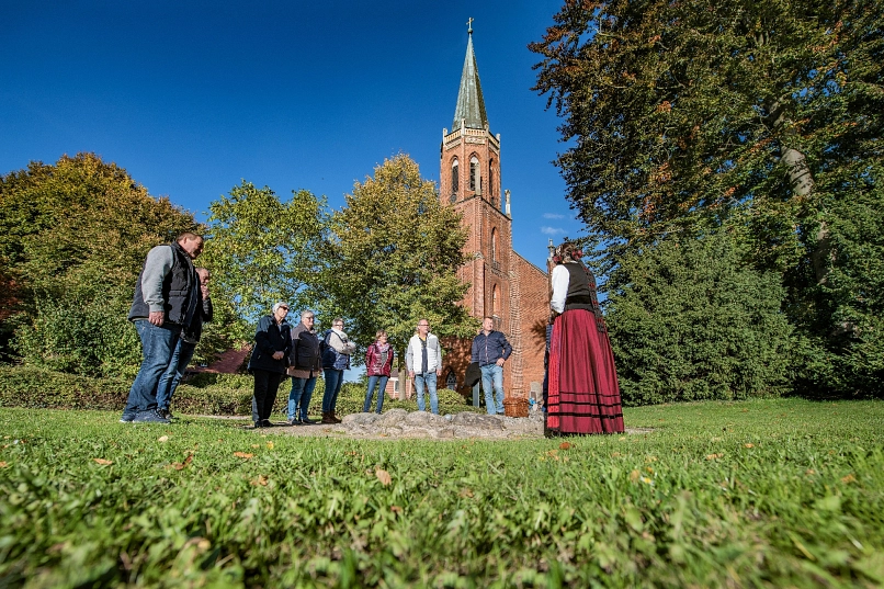Gruppe im Kreis stehender Menschen vor Dame in historischer Tracht, die von hinten zu sehen ist, im Hintergrund ein hoher Kirchturm