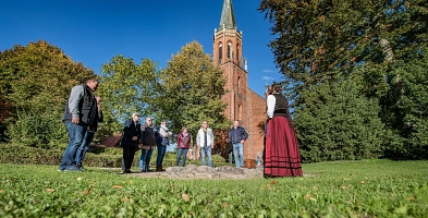 Gruppe im Kreis stehender Menschen vor Dame in historischer Tracht, die von hinten zu sehen ist, im Hintergrund ein hoher Kirchturm
