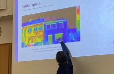 Vortrag Thermografiespaziergang Auf einer Leinwand ist ein Haus mit den bunten Bildern einer Wärmebildkamera zu sehen. Davor steht eine Person, die sich zu dem Bild dreht und etwas darauf zeigt.